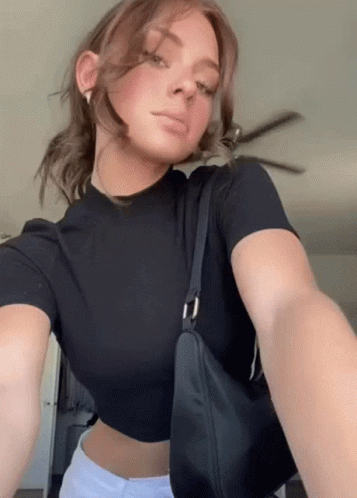 A Woman In A Black Crop Top Is Taking A Selfie While Holding A Black Bag . GIF