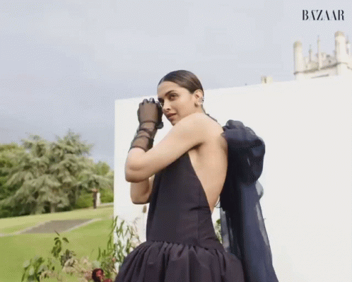 A Woman In A Black Dress And Black Gloves Is Standing In Front Of A White Wall That Says Bazaar On It GIF