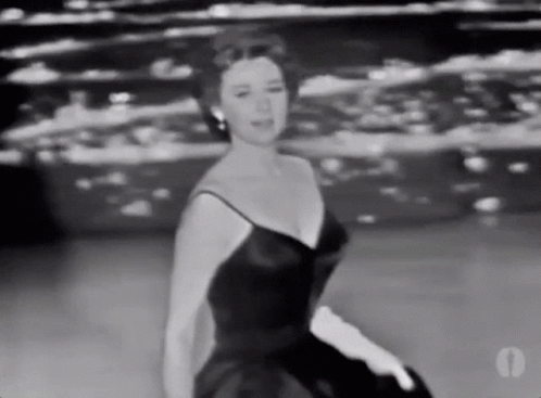 A Woman In A Black Dress And White Gloves Is Dancing On A Stage . GIF