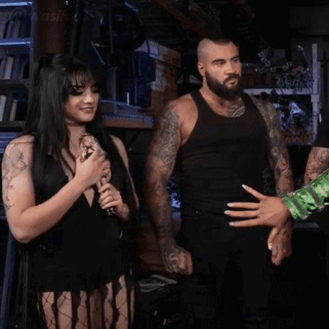 A Woman In A Black Dress Is Holding A Microphone And Smiling While A Man Stands Behind Her . GIF