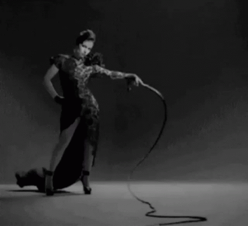 A Woman In A Black Dress Is Holding A Whip In Her Hand GIF