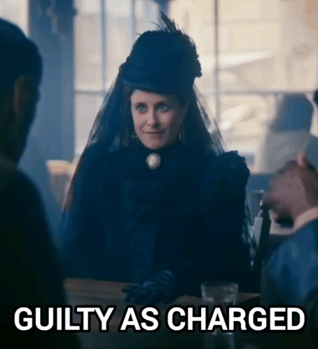 A Woman In A Black Dress Is Sitting At A Table With A Sign That Says Guilty As Charged GIF