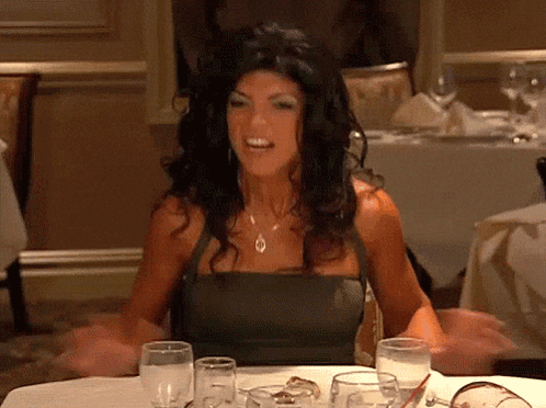 A Woman In A Black Dress Is Sitting At A Table With Wine Glasses GIF