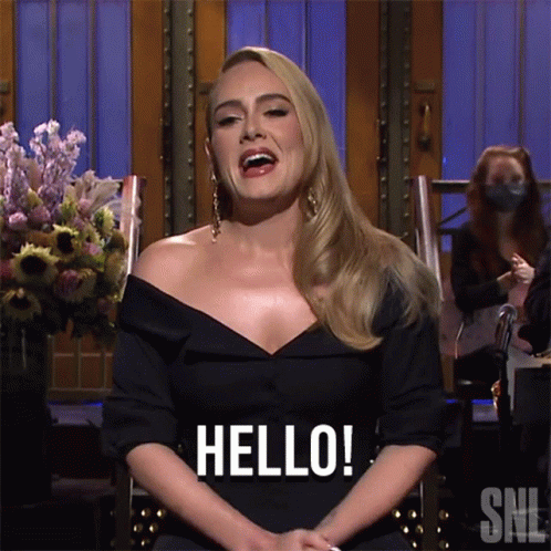A Woman In A Black Dress Is Sitting In A Chair And Saying Hello GIF