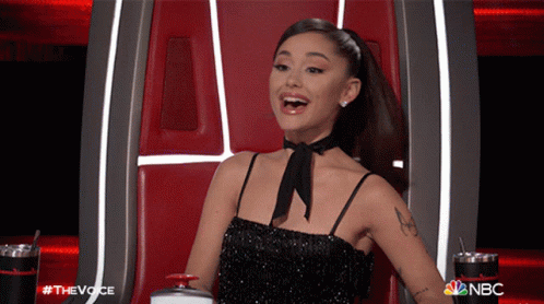 A Woman In A Black Dress Is Sitting In A Chair With The Nbc Logo Behind Her GIF