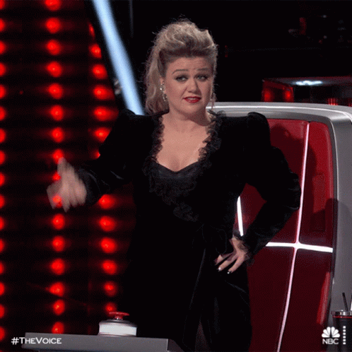 A Woman In A Black Dress Is Sitting In Front Of A Nbc Sign GIF