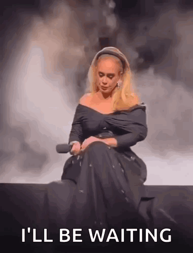 A Woman In A Black Dress Is Sitting On A Stage Holding A Microphone And The Words `` I 'Ll Be Waiting '' GIF