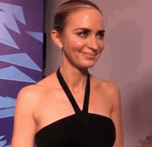 A Woman In A Black Dress Is Smiling In Front Of A Purple Wall . GIF