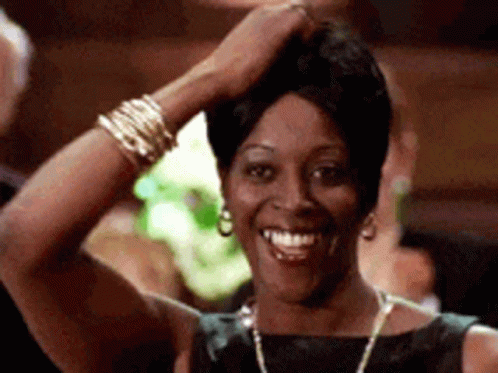 A Woman In A Black Dress Is Smiling With Her Hand On Her Head . GIF