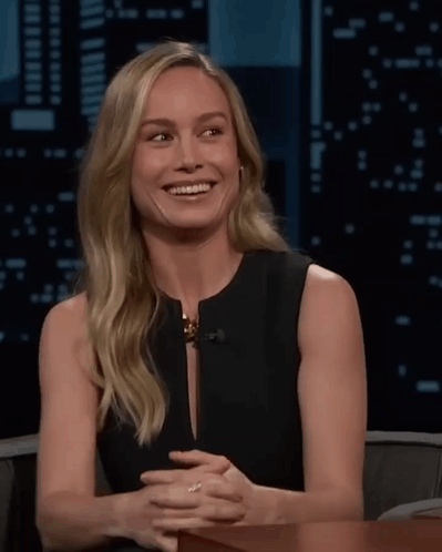 A Woman In A Black Dress Is Smiling With Her Hands Folded In Front Of Her GIF