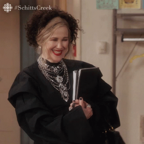A Woman In A Black Dress Is Smiling With #Schitts Creek On The Bottom Right GIF