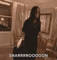 A Woman In A Black Dress Is Standing In A Living Room Holding A Bag And A Purse . GIF