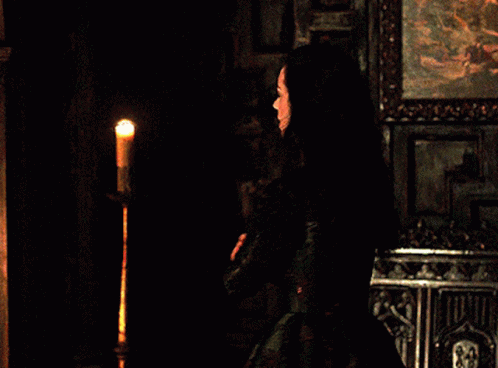 A Woman In A Black Dress Is Standing In Front Of A Candle GIF