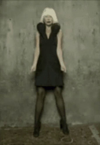 A Woman In A Black Dress Is Standing In Front Of A Gray Wall GIF