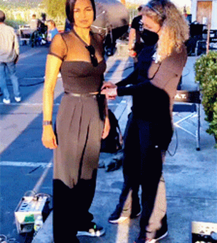 A Woman In A Black Dress Is Standing Next To A Woman In A Black Shirt With A T On It GIF