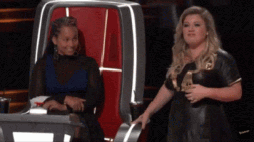 A Woman In A Black Dress Is Standing Next To Another Woman In A Chair GIF