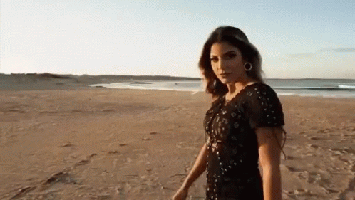 A Woman In A Black Dress Is Standing On A Sandy Beach GIF