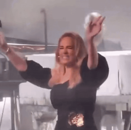 A Woman In A Black Dress Is Standing On A Stage With Her Arms In The Air . GIF