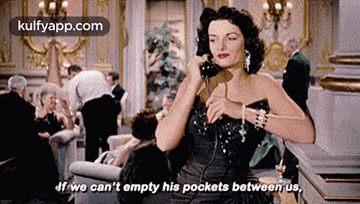 A Woman In A Black Dress Is Talking On A Phone In A Room . GIF