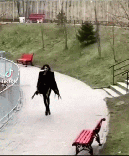 A Woman In A Black Dress Is Walking Down A Path With A Red Bench In The Background . GIF