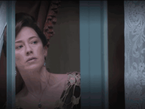 A Woman In A Black Dress Looks Out A Window With A Blue Curtain Behind Her GIF