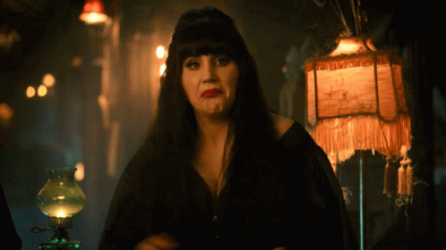 A Woman In A Black Dress Stands In Front Of A Lamp With A Fringed Shade That Says ' A ' GIF