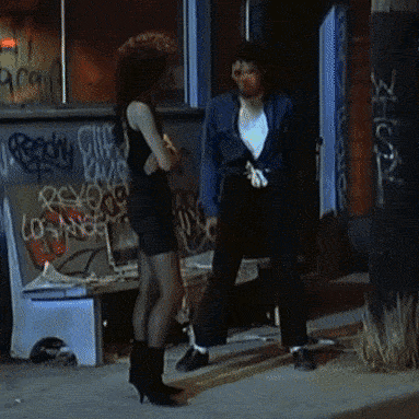 A Woman In A Black Dress Stands Next To A Man In A Blue Jacket In Front Of A Building With Graffiti On It GIF