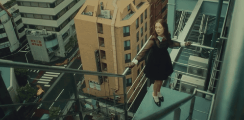 A Woman In A Black Dress Stands On A Balcony Overlooking A City With A Building That Says Comic Shop On It GIF