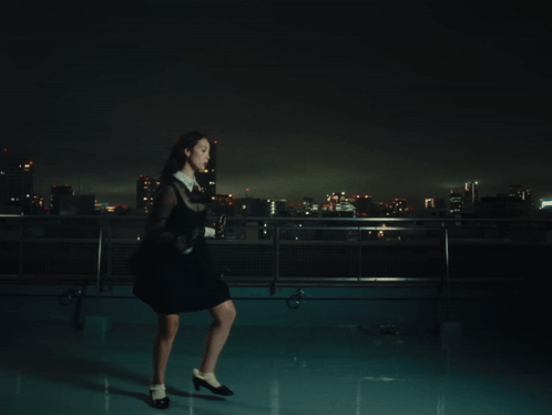 A Woman In A Black Dress Stands On A Rooftop At Night GIF