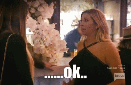 A Woman In A Black Dress Talks To Another Woman In Front Of A Sign That Says Ok On It GIF