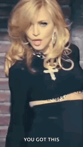 A Woman In A Black Dress With A Cross Necklace Is Standing In Front Of A Brick Wall And Says You Got This . GIF