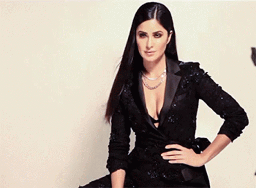 A Woman In A Black Dress With A Plunging Neckline Is Sitting On A Chair . GIF