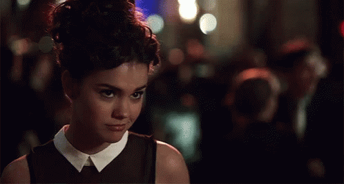 A Woman In A Black Dress With A White Collar Looks At The Camera GIF