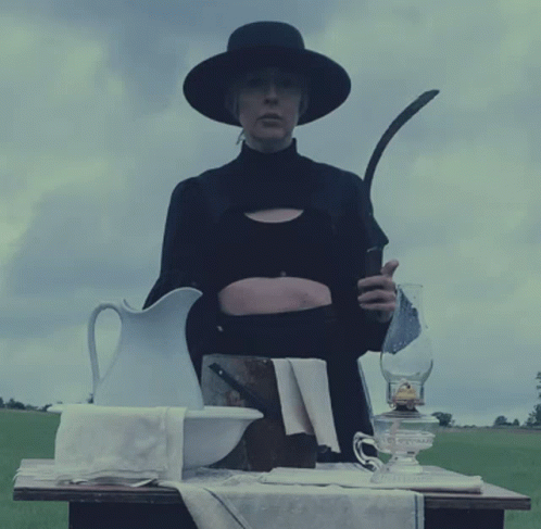 A Woman In A Black Hat Holds A Sickle In Her Hand GIF