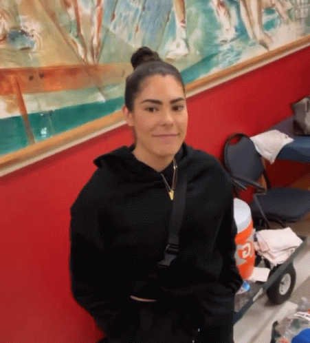 A Woman In A Black Hoodie Is Standing In Front Of A Red Wall And Smiling . GIF