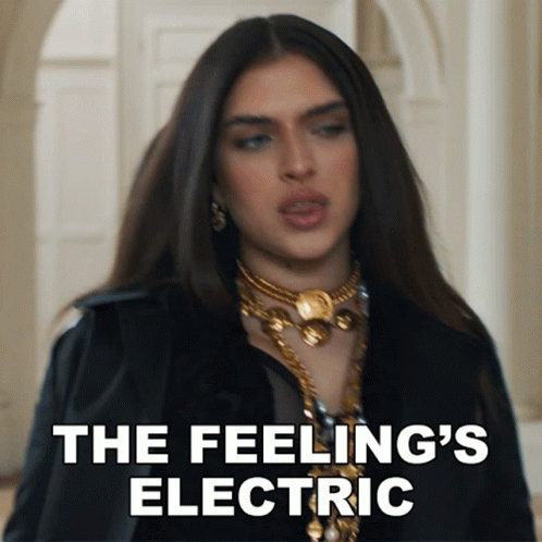 A Woman In A Black Jacket And Gold Necklace Says " The Feeling 'S Electric " In White Letters GIF