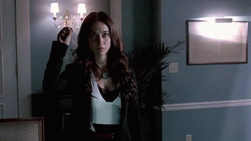 A Woman In A Black Jacket And White Top Stands In A Dark Room GIF