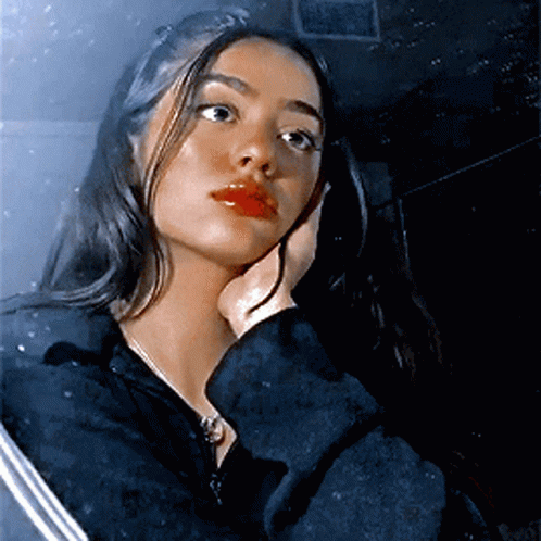 A Woman In A Black Jacket Holds Her Hand To Her Face And Looks At The Camera GIF