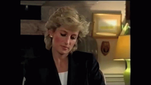 A Woman In A Black Jacket Is Sitting At A Table With A Lamp In The Background . GIF