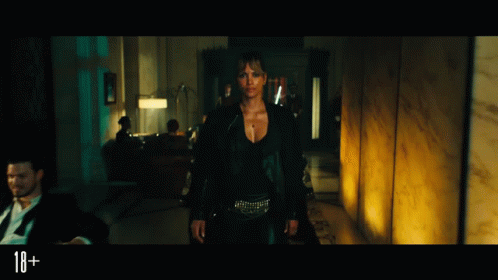 a woman in a black jacket is standing in a dark room with a sign that says 18+ in the corner