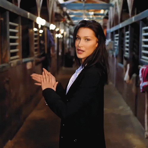 A Woman In A Black Jacket Is Standing In A Stable With Her Hands Folded GIF