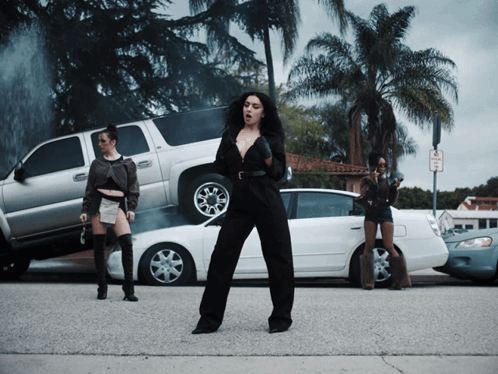 A Woman In A Black Jumpsuit Is Standing In Front Of A Stack Of Cars And A No Parking Sign GIF