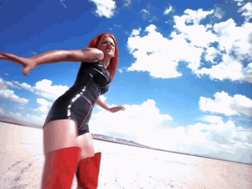 A Woman In A Black Latex Dress And Red Boots Is Standing On A Beach GIF