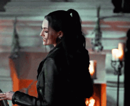 A Woman In A Black Leather Jacket Is Smiling In Front Of Candles GIF