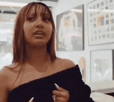 A Woman In A Black Off The Shoulder Top Is Standing In Front Of A Wall With Paintings On It . GIF