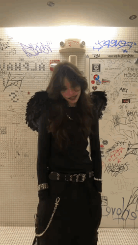 A Woman In A Black Outfit Is Standing In Front Of A Wall That Has Graffiti On It Including The Word Ovoks GIF