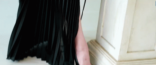 A Woman In A Black Pleated Skirt Is Standing Next To A White Column GIF
