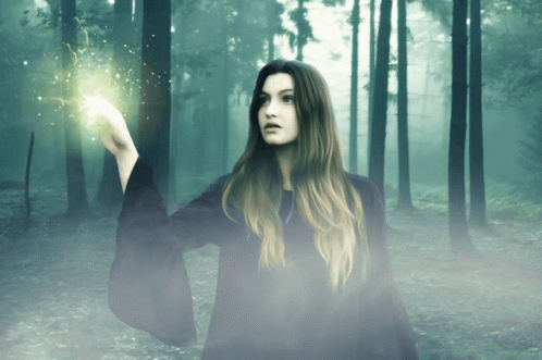 A Woman In A Black Robe Is Holding A Light In Her Hand In A Forest GIF