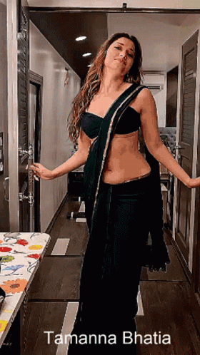 A Woman In A Black Saree Is Standing In A Hallway With The Name Tamanna Bhatia On The Bottom Right GIF