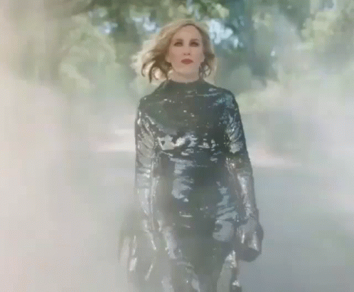 A Woman In A Black Sequined Jumpsuit Is Walking Through A Foggy Forest . GIF